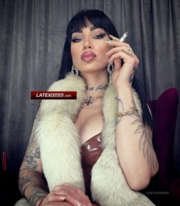 Inked Mistress with black hair smoking, wearing white fur stole and brown latex top