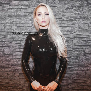 Just Lucy posing in a shiny black latex catsuit against a gray stone wall.