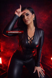Mistress in a black wet-look catsuit making a loser sign gesture against a red smoky background.