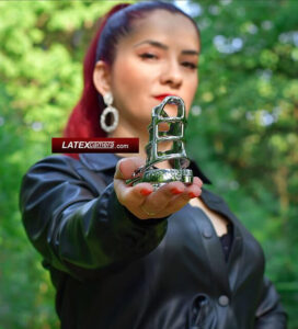 Red-haired Mistress in a black leather coat holding a metal penis cage outdoors.