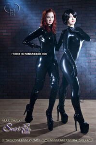 Two fetish models wearing black latex catsuits, black latex gloves, and black PVC platform boots posing against a dark brick wall.