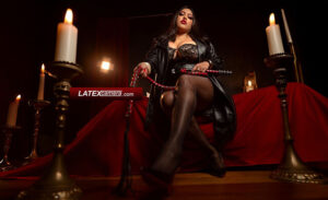 BBW Dominatrix wearing a black leather coat, nylon pantyhose, and high heels while holding a red whip in a candlelit setting.