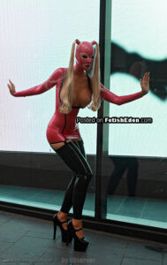 Fetish model wearing a red latex mini-dress, matching latex hood, black latex thigh-high stockings, and platform high heels near a glass wall.