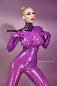 Miss Nyu posing in a shiny translucent purple latex catsuit with matching latex gloves and a purple neck corset against a purple studio background.