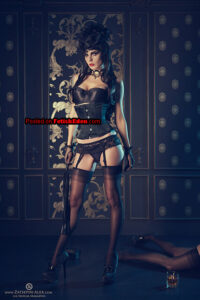 Mistress Elisanth wearing a black corset, lingerie, nylon stockings, garters, high heels, and gloves while holding a flogger in an elegant dark interior.