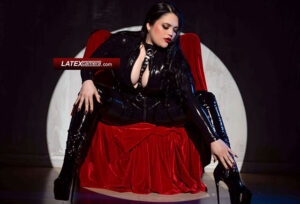 Voluptuous Domme wearing a black PVC catsuit and black PVC over-the-knee boots while seated in a chair.