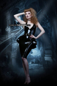 Blonde fetish model wearing a black sleeveless latex midi-dress with a belt, peplum details, black high heels, and a studded choker while posing in front of a gothic architectural backdrop.