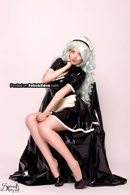 Fetish model wearing a black latex maid mini-dress with white trim, matching headpiece, platinum wavy hair, and black high heels while posing on a chair.